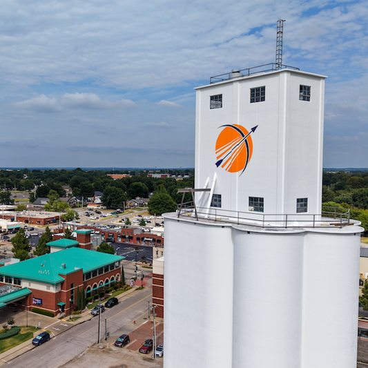 Aerial view of a large white cylindrical silo with a white tower section, featuring a prominent orange circular logo with a white arrow on the side. The silo is situated in an industrial urban area with roads, parking lots, and surrounding buildings under a blue sky with scattered clouds.